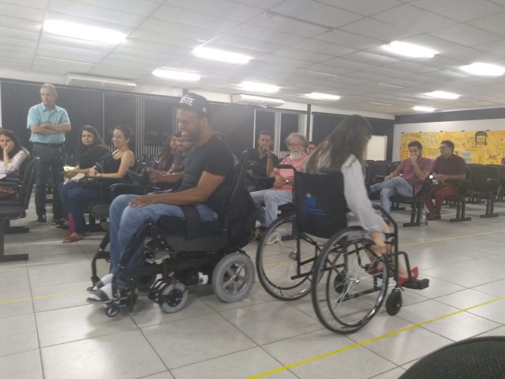 #pratodosverem imagem que registra o momento de uma dinâmica realizada no auditório. Ao centro um homem e uma mulher, de costas um para o outro, ambos, em cadeiras de rodas. À direita pessoas na plateia observam a atividade. Fim da descrição.